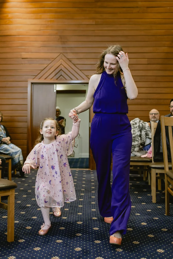 A young flower girl in a pink dress and a bridesmaid in a vibrant purple jumpsuit walking down the aisle, captured during a beautiful celebration of small wedding photography in Southampton by VasPhotography