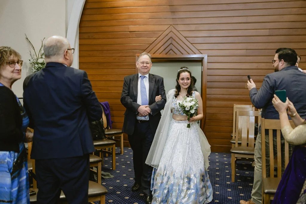 A radiant bride in a white and blue patterned lehenga walking down the aisle with her father, an example of stunning small wedding photography in Southampton by VasPhotography.