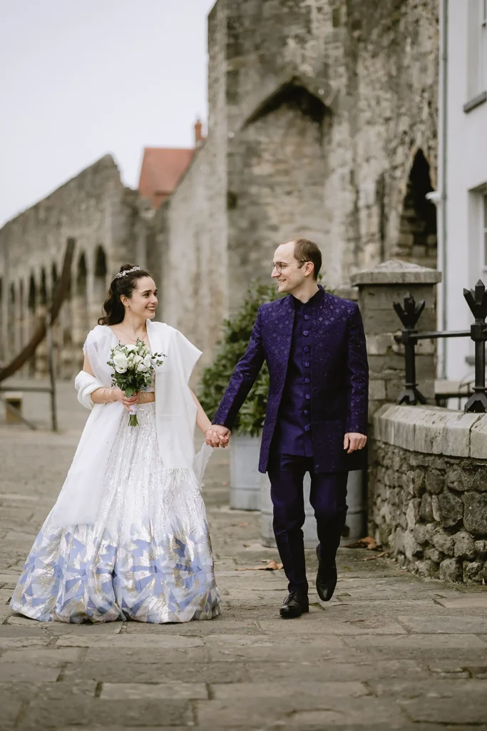 A newly married bride and groom holding hands, smiling, and posing for portraits outdoors against scenic stone walls and garden pathways, captured beautifully by VasPhotography specializing in small wedding photography in Southampton