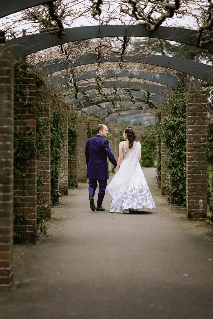A newly married bride and groom holding hands, smiling, and posing for portraits outdoors against scenic stone walls and garden pathways, captured beautifully by VasPhotography specializing in small wedding photography in Southampton