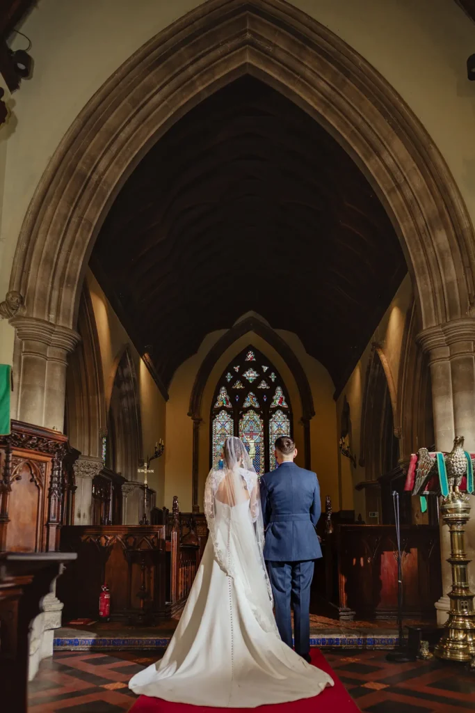 Candid wedding photographer capturing bride and groom during church ceremony with stained glass backdrop.