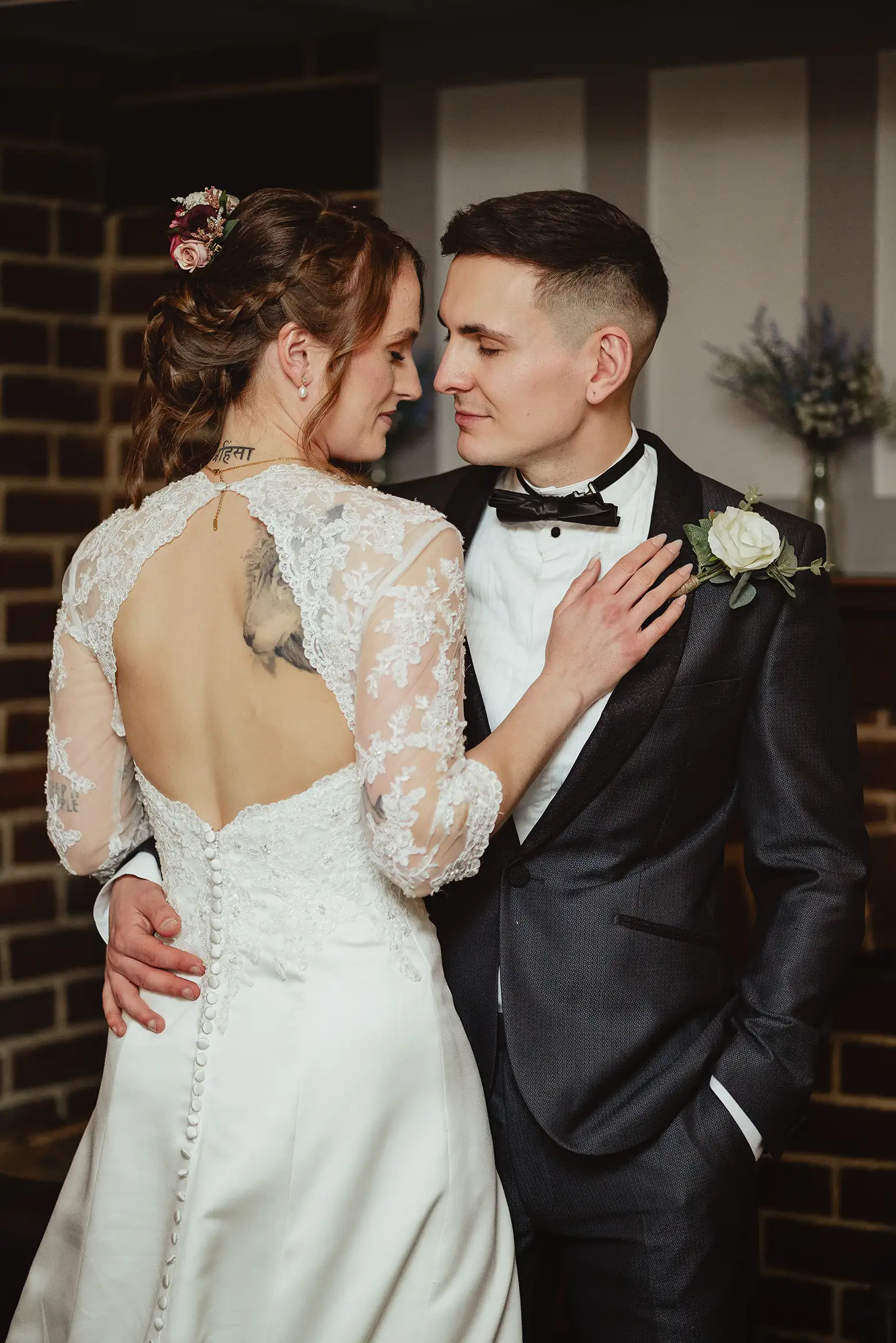 Candid wedding photographer capturing intimate bride and groom portrait with lace back dress and black tuxedo indoors.