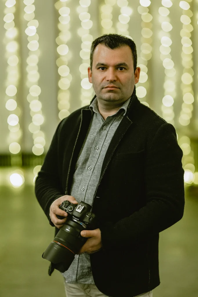 Vas from VasPhotography holding a professional camera in front of blurred string lights